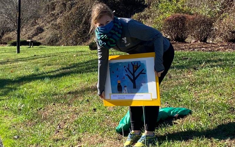 Williams YMCA Mitchell County Community Liaison Katie Willett smiles under her face covering as she places a sign in the ground advertising the new story trail in Riverside Park. The pilot story for the trail is called “Wonderfall” and follows the journey of a tree as it changes throughout the four seasons. The story mascot, Piney the Squirrel, is also featured prominently on the trail and is available for pictures with visitors. Piney was designed by Day Dotson and the trail project is a collaboration betw
